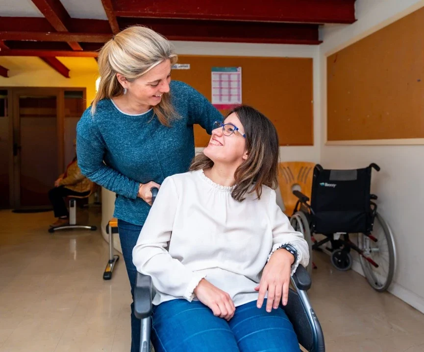A disabled woman smiling while sitting in her wheelchair and her support worker